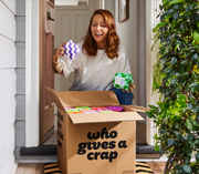 A woman smiling and holding up two packaged items while standing in a doorway with a large cardboard box labeled 'who gives a crap' in front of her. The box is filled with colorful packaged goods, and a green potted plant is visible on the right.