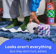 A person in striped pajamas and green socks rolls a toilet paper tube on a blue shag rug, with a stack of colorful patterned toilet paper tubes standing nearby. A blue text banner at the bottom reads, 'Looks aren't everything (but they don't hurt, either)'.