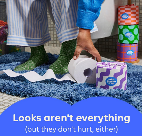 A person in striped pajamas and green socks rolls a toilet paper tube on a blue shag rug, with a stack of colorful patterned toilet paper tubes standing nearby. A blue text banner at the bottom reads, 'Looks aren't everything (but they don't hurt, either)'.